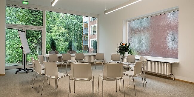 Heller Tagungsraum mit Stuhlkreis und Präsentationstechnik. Durch die großen Fenster fällt Tageslicht in den Raum und der Blick geht ins Grüne.