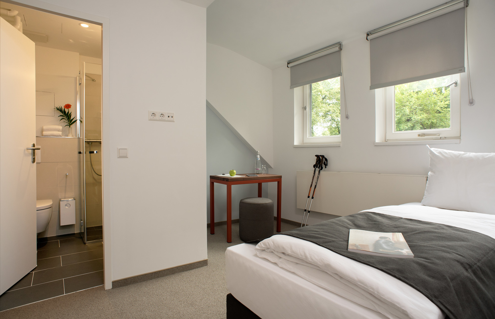Helles Hotelzimmer mit Doppelbett und zwei kleinen Fenstern. Links ist der offene Zugang zum Badezimmer zu sehen.